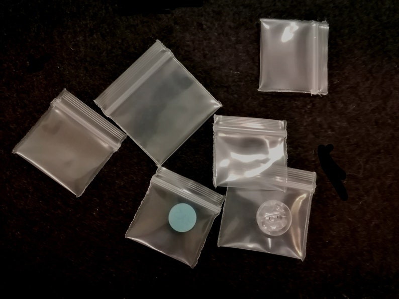 May include: Six clear plastic resealable bags with zipper closures. The bags are various sizes and are lying flat on a black surface. One bag has a small blue circle inside.