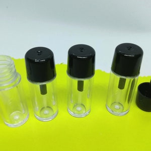 May include: Four clear glass bottles with black lids and a black applicator brush. The bottles are empty and are arranged in a row on a yellow surface.