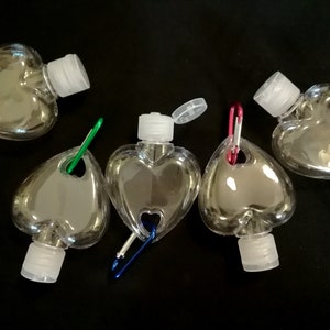 May include: Five clear plastic heart-shaped bottles with attached carabiners. The bottles have a small opening at the top with a white cap. The carabiners are different colors: purple, green, blue, red, and red.