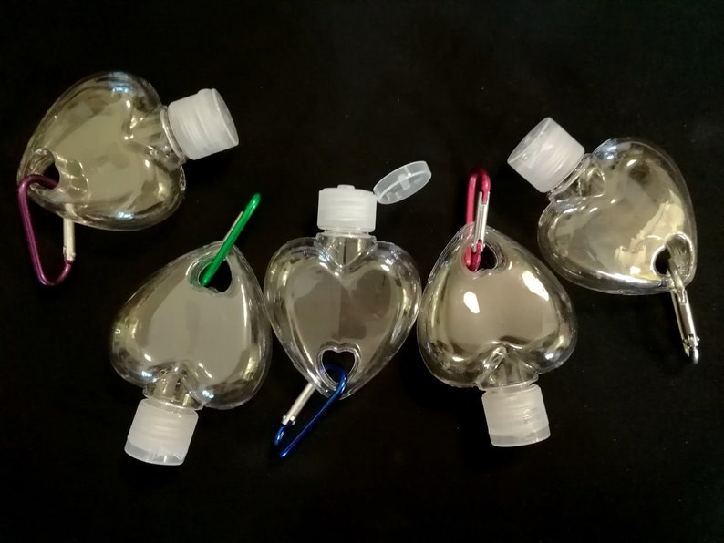 May include: Five clear plastic heart-shaped bottles with attached carabiners. The bottles have a small opening at the top with a white cap. The carabiners are different colors: purple, green, blue, red, and red.