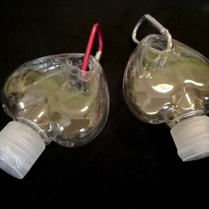 May include: Two clear plastic heart-shaped bottles with silver carabiner clips. The bottles have white screw-on caps.