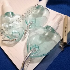 May include: Three heart-shaped, clear plastic spray bottles with white spray nozzles and silver carabiner clips. The bottles are on a spiral notebook with a wooden pencil that has the text "www.IKEA.com".
