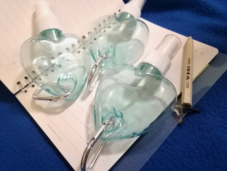 May include: Three heart-shaped, clear plastic spray bottles with white spray nozzles and silver carabiner clips. The bottles are on a spiral notebook with a wooden pencil that has the text "www.IKEA.com".