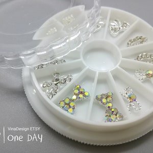 Könnte beinhalten: Ein weißes Plastikrad mit 12 Fächern, die jeweils ein anderes Design aus silbernen und schillernden Strasssteinen enthalten. Das Rad ist offen und der Deckel liegt daneben.