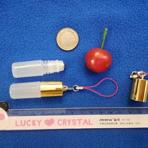 Op de afbeelding: Twee transparante glazen rollerflessen met gouden doppen. E&eacute;n fles is bevestigd aan een roze koord met een gouden hanger. Een liniaal met de tekst "LUCKY CRYSTAL" is zichtbaar in de onderkant van de afbeelding.