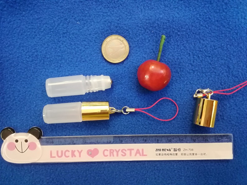 Op de afbeelding: Twee transparante glazen rollerflessen met gouden doppen. E&eacute;n fles is bevestigd aan een roze koord met een gouden hanger. Een liniaal met de tekst "LUCKY CRYSTAL" is zichtbaar in de onderkant van de afbeelding.