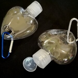 May include: Two clear plastic heart-shaped bottles with white screw-on caps and a silver carabiner clip attached to one bottle.