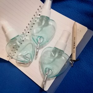 May include: Three heart-shaped, clear plastic spray bottles with white spray nozzles. Each bottle has a silver-colored clip attached. The bottles are on a spiral-bound notebook and a blue surface. A pencil with the text "www. IKEA .com" is also present.