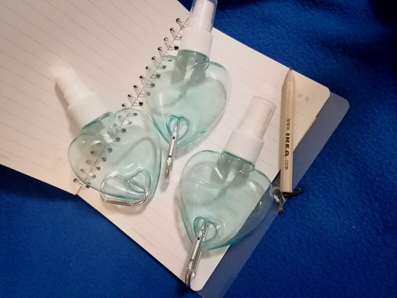 May include: Three heart-shaped, clear plastic spray bottles with white spray nozzles. Each bottle has a silver-colored clip attached. The bottles are on a spiral-bound notebook and a blue surface. A pencil with the text "www. IKEA .com" is also present.