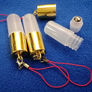 Op de afbeelding: Twee transparante glazen rollerflessen met gouden doppen en rode koordjes. Een derde transparante glazen rollerfles met een zilveren rolbal is ook te zien.