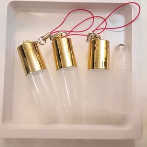Op de afbeelding: Vier transparante glazen rollerball-flesjes met gouden dopjes en een rood touwtje dat aan de dopjes is bevestigd. De flesjes staan op een wit oppervlak.