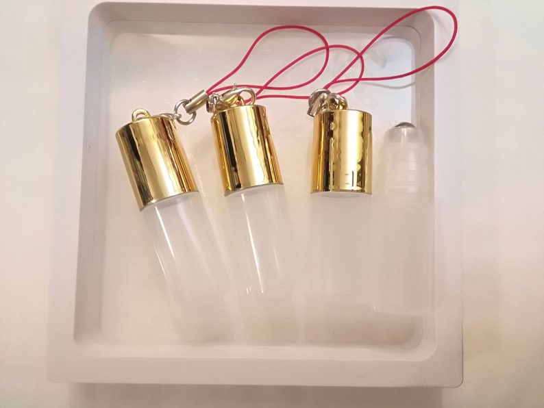 Op de afbeelding: Vier transparante glazen rollerball-flesjes met gouden dopjes en een rood touwtje dat aan de dopjes is bevestigd. De flesjes staan op een wit oppervlak.