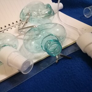 May include: Clear, oval-shaped plastic spray bottles with white spray nozzles and a clear cap. The bottles are partially disassembled, with some components lying on a notebook. The background is a blue surface.