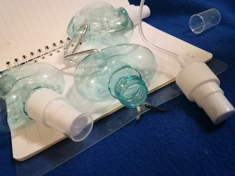 May include: Clear, oval-shaped plastic spray bottles with white spray nozzles and a clear cap. The bottles are partially disassembled, with some components lying on a notebook. The background is a blue surface.