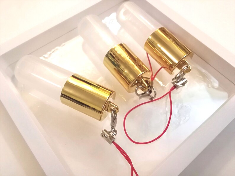 Op de afbeelding: Drie transparante glazen flessen met gouden doppen en een rood touwtje eraan vastgemaakt. De flessen bevinden zich in een witte doos.