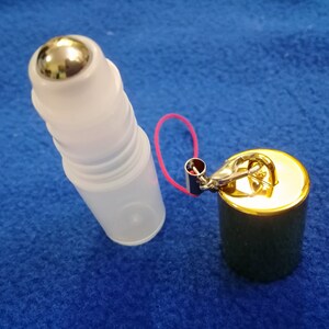 Op de afbeelding: Een transparante glazen rollerfles met een zilveren metalen rollerbal, bevestigd aan een gouden metalen dop met een klein rood koord en een gouden metalen sluiting.