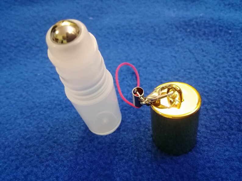 Op de afbeelding: Een transparante glazen rollerfles met een zilveren metalen rollerbal, bevestigd aan een gouden metalen dop met een klein rood koord en een gouden metalen sluiting.