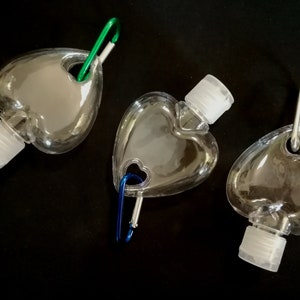 May include: Three clear plastic heart-shaped bottles with silver caps and a carabiner clip attached to each. The bottles are empty and appear to be for hand sanitizer.