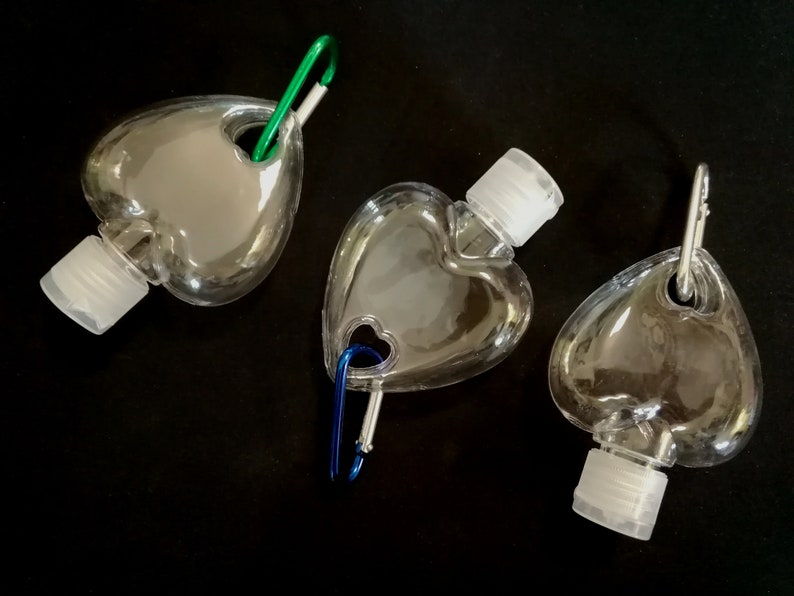 May include: Three clear plastic heart-shaped bottles with silver caps and a carabiner clip attached to each. The bottles are empty and appear to be for hand sanitizer.