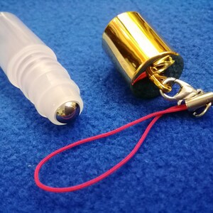 Op de afbeelding: Een transparante glazen rollerfles met een zilveren rollerbal bevestigd aan een gouden dop. De dop heeft een zilveren sluiting en een rood touwtje.