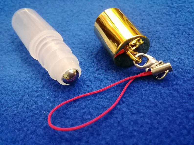 Op de afbeelding: Een transparante glazen rollerfles met een zilveren rollerbal bevestigd aan een gouden dop. De dop heeft een zilveren sluiting en een rood touwtje.