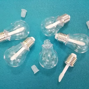 Puede incluir: Seis botellas transparentes de plástico con forma de bombilla, con tapas plateadas y aplicadores blancos. Las botellas están sobre una superficie azul.