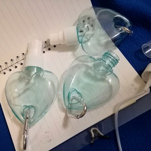 May include: Clear, heart-shaped spray bottles with white spray nozzles and silver carabiner clips. The bottles are partially disassembled, with one cap removed. The bottles are on a spiral-bound notebook.