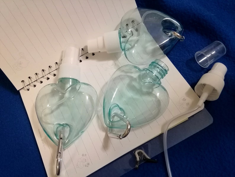 May include: Clear, heart-shaped spray bottles with white spray nozzles and silver carabiner clips. The bottles are partially disassembled, with one cap removed. The bottles are on a spiral-bound notebook.