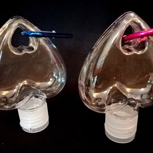 May include: Two clear plastic heart-shaped bottles with a small loop at the top. The bottles have white screw-on caps. One bottle has a blue loop and the other has a pink loop.