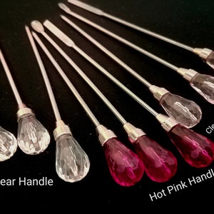 May include: Set of eight clear glass and hot pink glass stirring tools with silver handles. The tools have a teardrop-shaped tip.  The text "Clear Handle" and "Hot Pink Handle" are visible below the tools.  The text "Clear Light Pink" is visible above the tools.