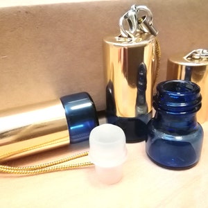 May include: Three small gold and blue glass bottles with a gold chain and a white plastic stopper. The bottles are on a light brown surface.