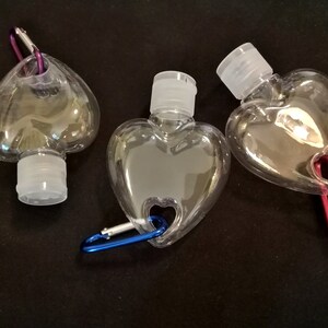 May include: Three clear plastic heart-shaped bottles with attached carabiners. The bottles have a small opening at the top with a white screw-on cap. The carabiners are different colors: purple, blue, and red.