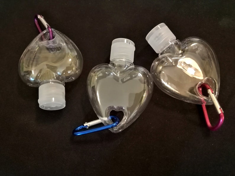 May include: Three clear plastic heart-shaped bottles with attached carabiners. The bottles have a small opening at the top with a white screw-on cap. The carabiners are different colors: purple, blue, and red.