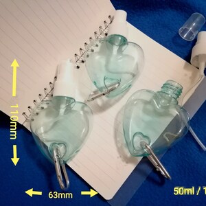 May include: Three heart-shaped, clear blue spray bottles with white spray nozzles and silver carabiner clips. Each bottle is approximately 4.3 inches tall and 2.5 inches wide, with a capacity of 1.7 ounces.
