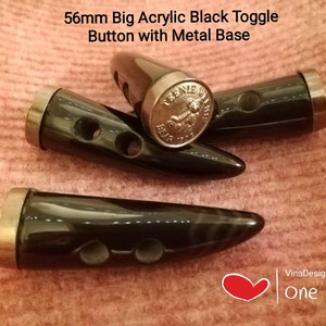 Puede incluir: Tres botones de palanca de acrílico negro con base de metal. Cada botón mide 56 mm de largo y tiene dos agujeros. Los botones están sobre un fondo de tela marrón.