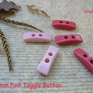 May include: Five pink and red plastic toggle buttons. The buttons are 35mm in size and have two holes each. The buttons are arranged in a row on a white fabric background.