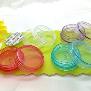 Może przedstawiać: Zestaw małych, przezroczystych pojemników plastikowych z pokrywkami w różnych kolorach, w tym czerwony, pomarańczowy, niebieski, fioletowy i przezroczysty. Pojemniki są ułożone jeden na drugim i znajdują się na zielonej powierzchni.