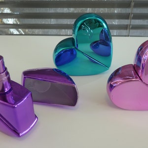 Puede incluir: Tres frascos de perfume en forma de corazón en diferentes colores: azul verdoso, rosa y morado. El frasco morado está abierto y tiene una boquilla de pulverización.