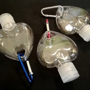 May include: Three clear heart-shaped plastic bottles with silver and blue carabiner clips. The bottles have white screw-on caps and are designed for carrying hand sanitizer.