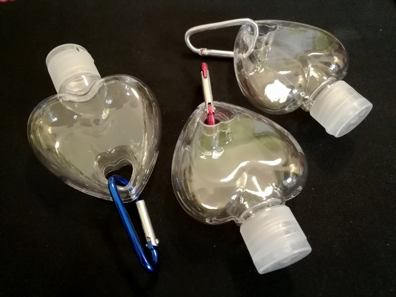 May include: Three clear heart-shaped plastic bottles with silver and blue carabiner clips. The bottles have white screw-on caps and are designed for carrying hand sanitizer.