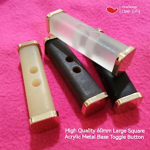 May include: Three large square toggle buttons with a 60mm acrylic base and gold metal accents. The buttons are in white, black, and clear.