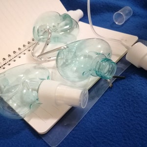 May include: Clear, heart-shaped spray bottles with white spray nozzles and silver carabiner clips. The bottles are on a white spiral notebook and a blue surface. A clear tube and a small clear cylinder are also present.