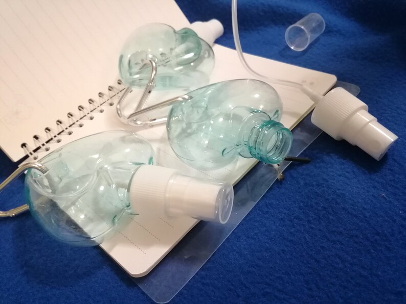 May include: Clear, heart-shaped spray bottles with white spray nozzles and silver carabiner clips. The bottles are on a white spiral notebook and a blue surface. A clear tube and a small clear cylinder are also present.