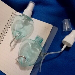 May include: Two heart-shaped, clear plastic spray bottles with white spray nozzles and silver carabiner clips. One bottle is open, revealing the interior. The bottles rest on a spiral-bound notebook.