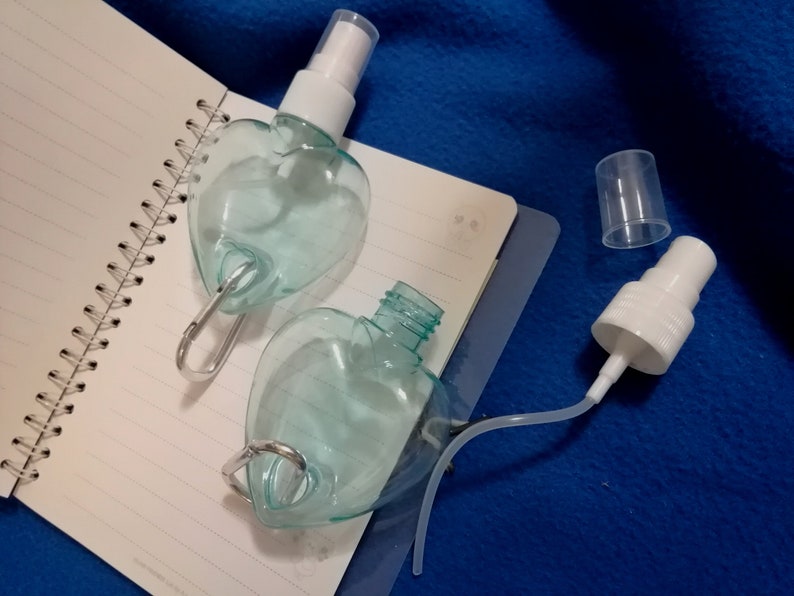 May include: Two heart-shaped, clear plastic spray bottles with white spray nozzles and silver carabiner clips. One bottle is open, revealing the interior. The bottles rest on a spiral-bound notebook.