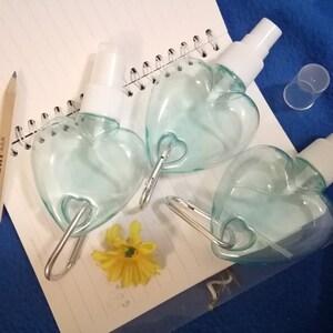 May include: Three heart-shaped, clear blue spray bottles with white spray nozzles and silver carabiner clips. A yellow flower and a pencil are on a spiral notebook. The notebook is on a blue surface.