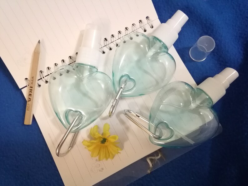 May include: Three heart-shaped, clear blue spray bottles with white spray nozzles and silver carabiner clips. A yellow flower and a pencil are on a spiral notebook. The notebook is on a blue surface.