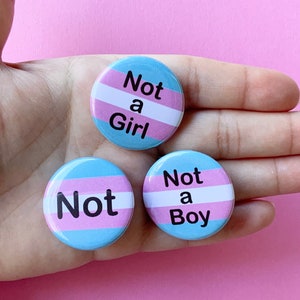 May include: Three round buttons with the transgender pride flag colors. The buttons say "Not a Girl", "Not a Boy", and "Not" in black text.