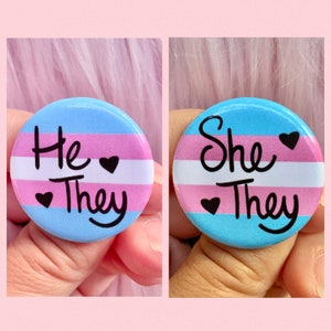 May include: Two round buttons with the transgender pride flag colors. The first button says "He/They" with a heart symbol between the words. The second button says "She/They" with a heart symbol between the words.
