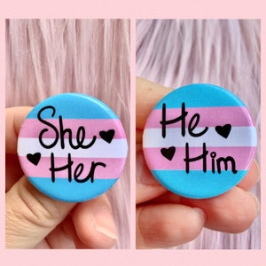 May include: Two round pins with the transgender pride flag colors. The left pin says "She Her" and the right pin says "He Him".